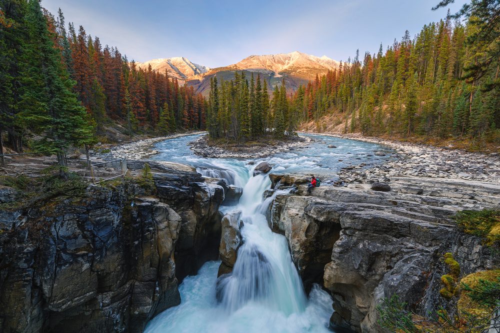 Sunwapta Falls mit Reisenden, die bei Sonnenuntergang auf einem Felsen im Herbstwald sitzen. Icefields Parkway, Jasper Nationalpark, Kanada