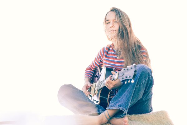 Caro Trischler sitzt mit einer Gitarre barfuß auf dem Boden