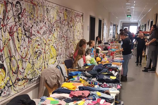 Kleider und Spielzeuge liegen auf langem Tisch und Menschen davor und dahinter