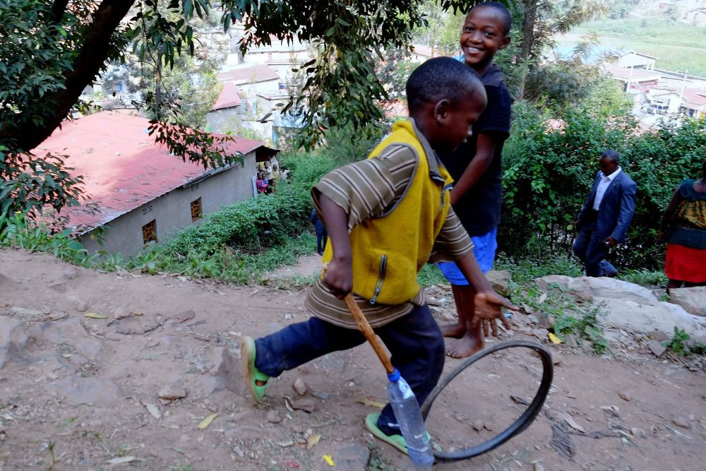 ein ruandesischer Junge treibt einen Reifen bergab, ein anderer schaut lachend zu
